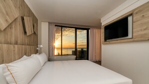 A modern hotel room with a large bed, a TV mounted on the wall, and a view of the sunset over the water. - Senator Gandia Hotel (Gandia)