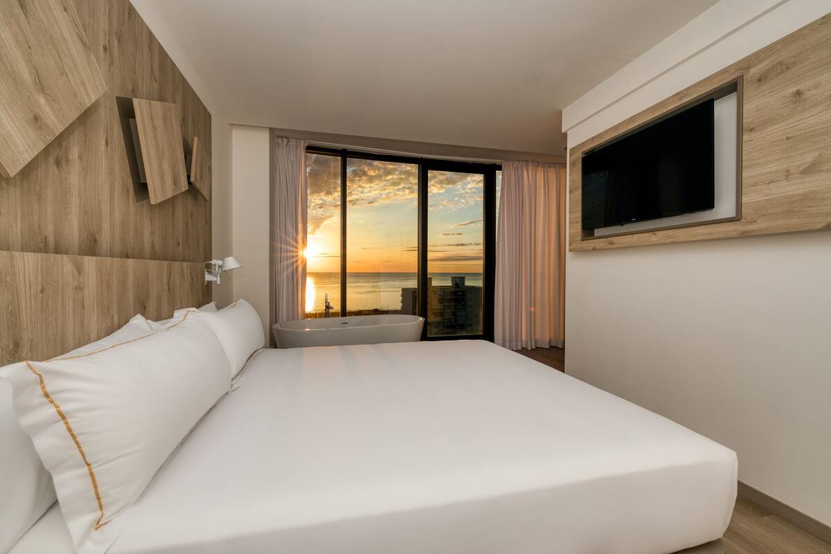 Une chambre d’hôtel moderne avec un grand lit, un téléviseur fixé au mur et une vue sur le coucher de soleil au-dessus de l’eau.