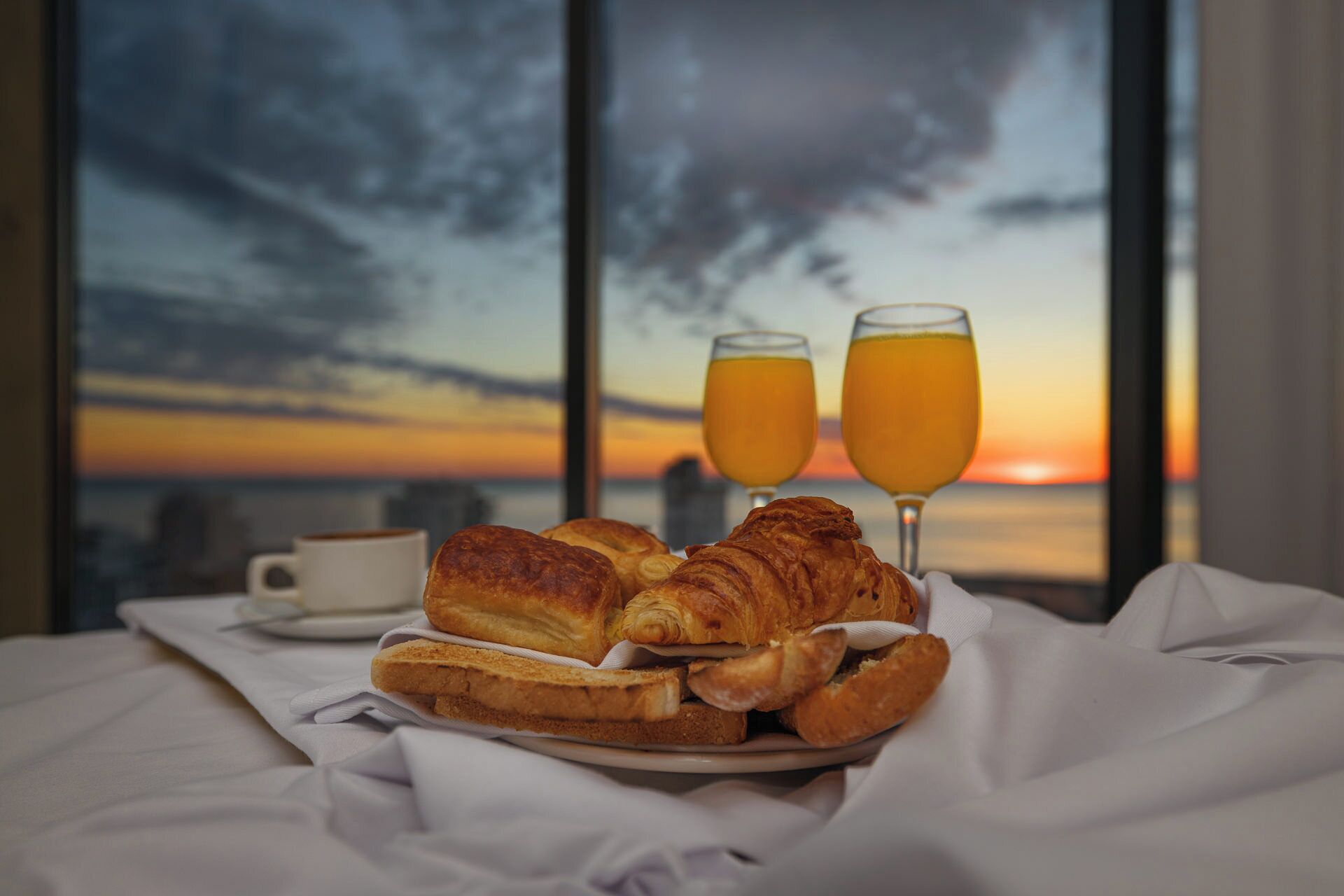 Desayuno con croissants, tostadas y jugo de naranja sobre una cama con vista a la ciudad al atardecer.