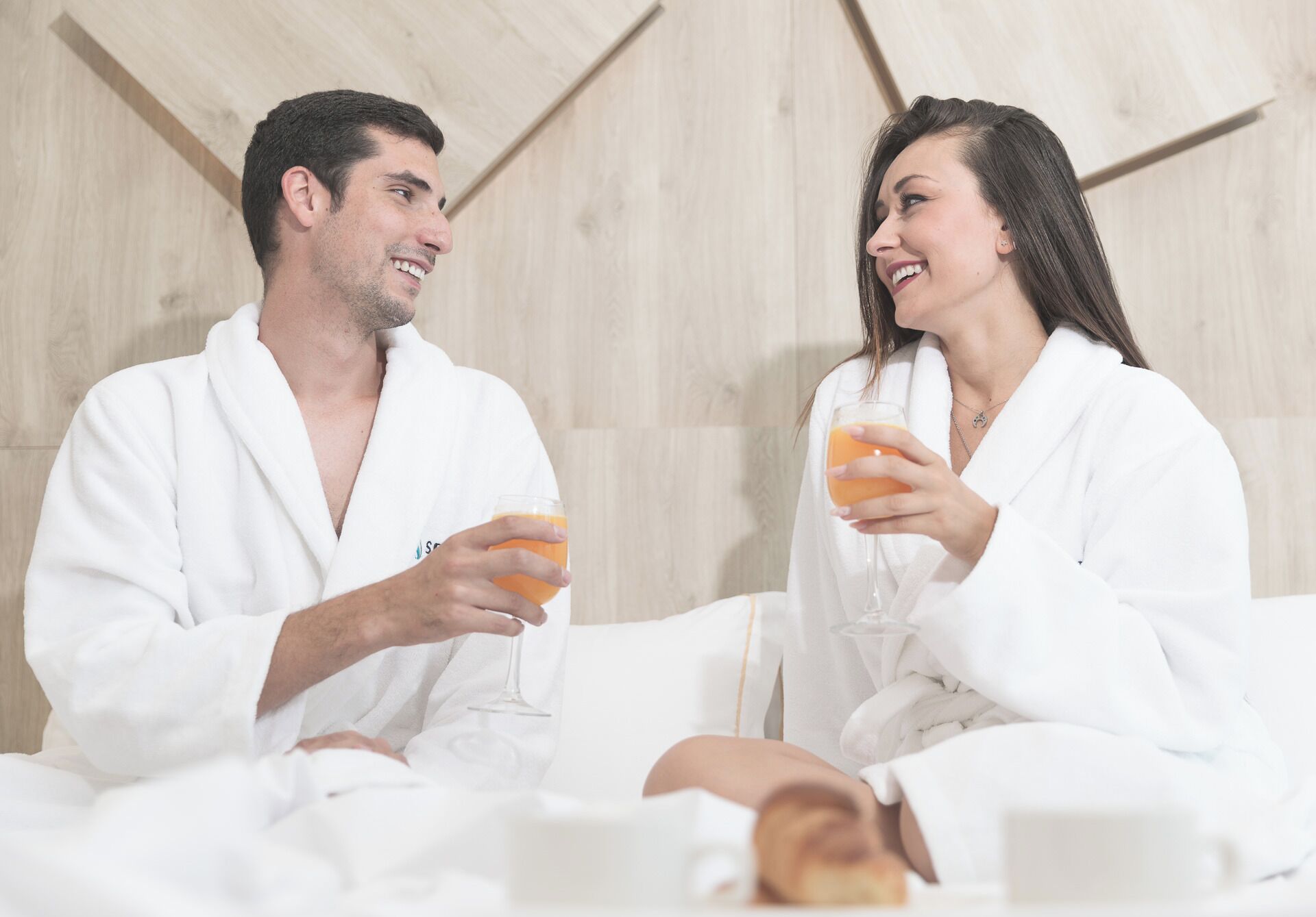 Two people in white bathrobes sitting on a bed with a wooden headboard, holding glasses of orange juice.