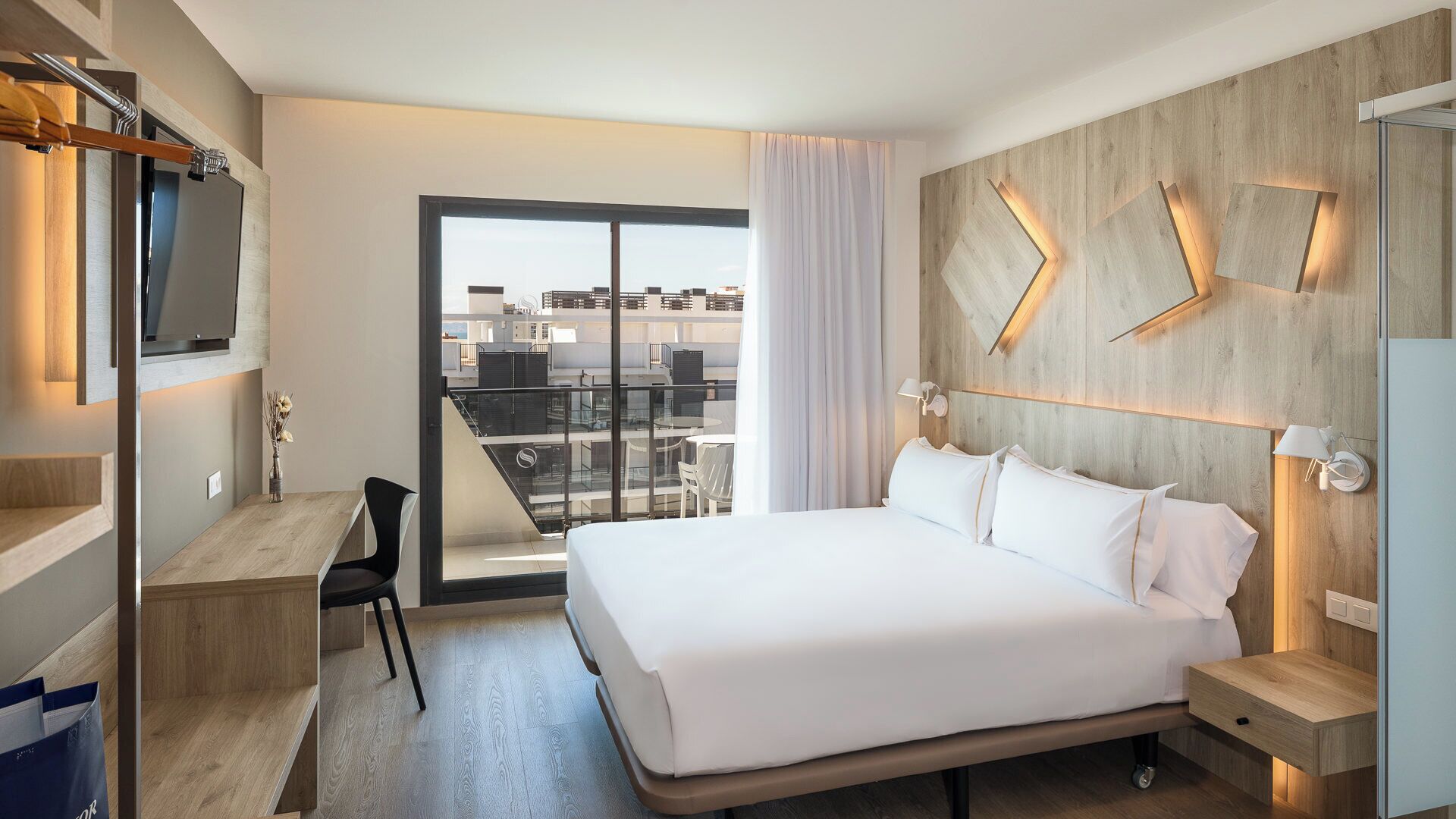 Habitación de hotel moderna con una cama grande, un escritorio, una silla y vistas a la ciudad a través de un amplio ventanal.