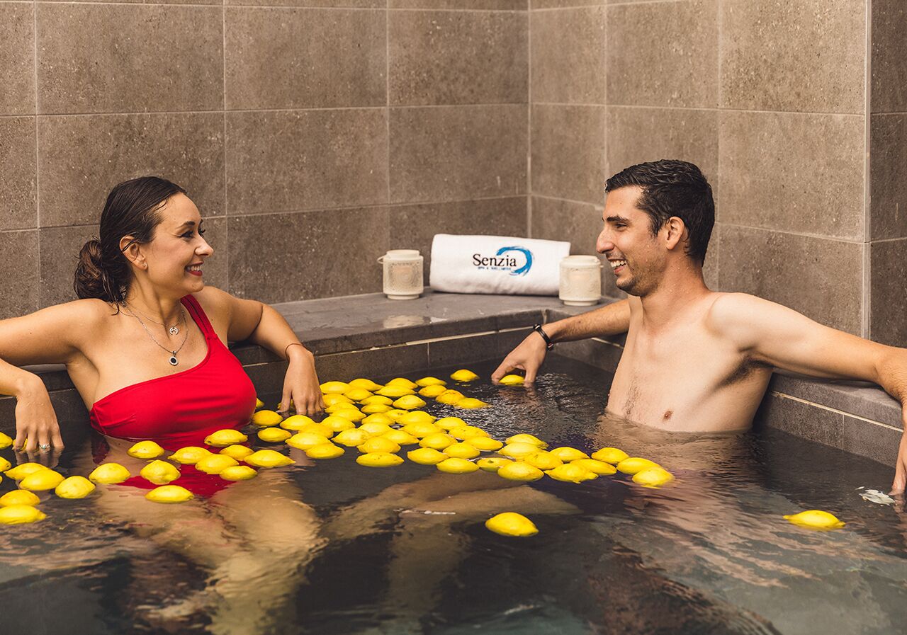 Una pareja en una bañera llena de limones, con una toalla y una taza a un lado.