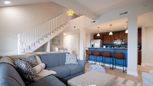 A modern living room with a grey sofa, patterned cushions, a wooden staircase, and a kitchen area with blue cabinets and bar stools. - Grand Haven Family Retreat | Game Loft • Sleeps 10 (Grand Haven)