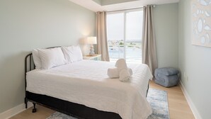 A bedroom with a bed, a nightstand, a lamp, a window with curtains, and a view of the outside. - Grand Haven Family Retreat | Game Loft • Sleeps 10 (Grand Haven)