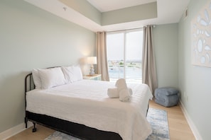 Un dormitorio con cama, mesita de noche, lámpara, ventana con cortinas y vista al exterior.