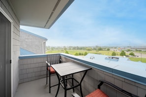 Un balcón con una mesa y sillas, vista de un paisaje verde y un edificio con techo azul.
