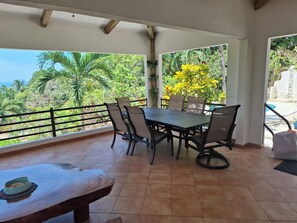 Une terrasse couverte, agrémentée d’une table et de chaises, entourée d’une balustrade, qui offre une vue sur un jardin luxuriant et sur la mer.