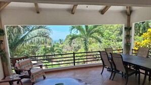 A covered patio with a wooden table and chairs, a bench with a striped cushion, and a view of lush greenery and palm trees. - Beautiful home with ocean views (Playa Samara, Nicoya)