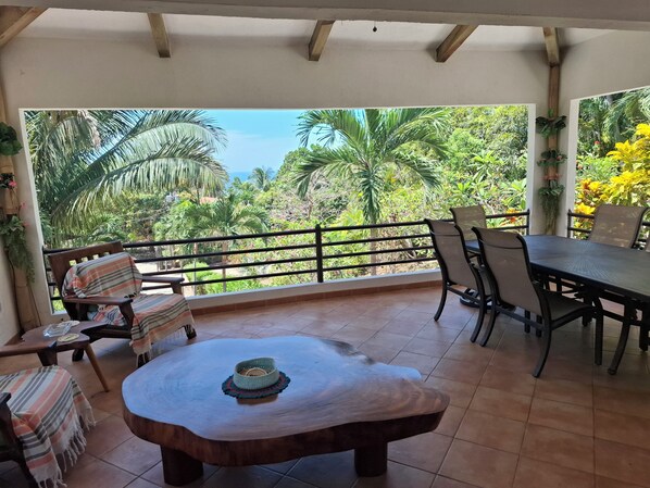 Une terrasse couverte avec une table et des chaises en bois, un banc agrémenté d’un coussin à rayures, et une vue sur une végétation luxuriante et des palmiers.