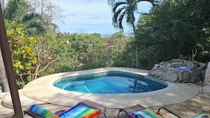 A swimming pool with a stone edge, surrounded by lush greenery and a view of the ocean. There are two lounge chairs with colorful towels. - Beautiful home with ocean views (Playa Samara, Nicoya)