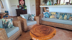 A living room with wicker furniture, a wooden coffee table, and a kitchen area with various bottles and plants. - Beautiful home with ocean views (Playa Samara, Nicoya)