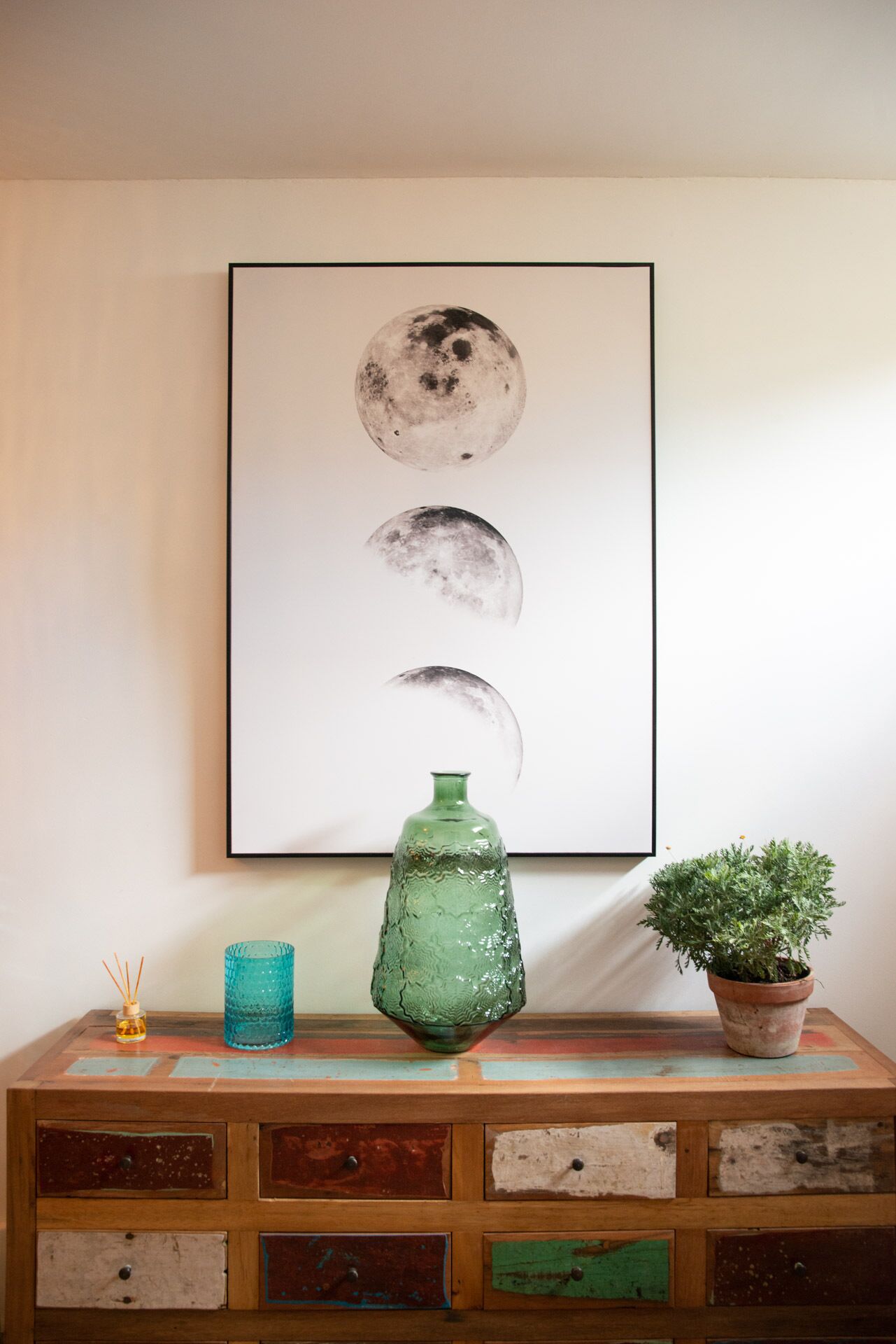 Une œuvre d’art représentant les différentes phases de la lune, fixée au mur, un vase en verre vert et une plante en pot, le tout posé sur une commode en bois dotée de tiroirs.