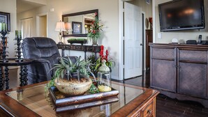 Un salón con una mesa de centro de vidrio, un sillón de cuero, un mueble de madera oscura y un televisor de pantalla plana montado en la pared.