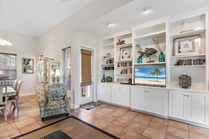 A living room with a patterned armchair, a dining table, and a bookshelf filled with books and decorative items.