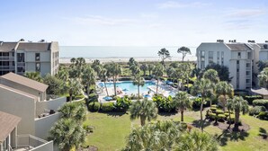Un resort con una piscina central rodeada de palmeras y sillas de descanso, con vista a una playa.