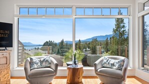 Amplia habitación con grandes ventanales que ofrecen vistas a la montaña, dos sillones grises, una mesa de centro de madera y un televisor sobre un mueble de madera.