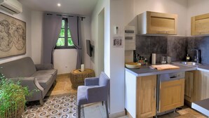 Un espacio habitable compacto con sofá, una mesita redonda, una silla, una zona de cocina con armarios de madera y una encimera, y una ventana con cortinas.