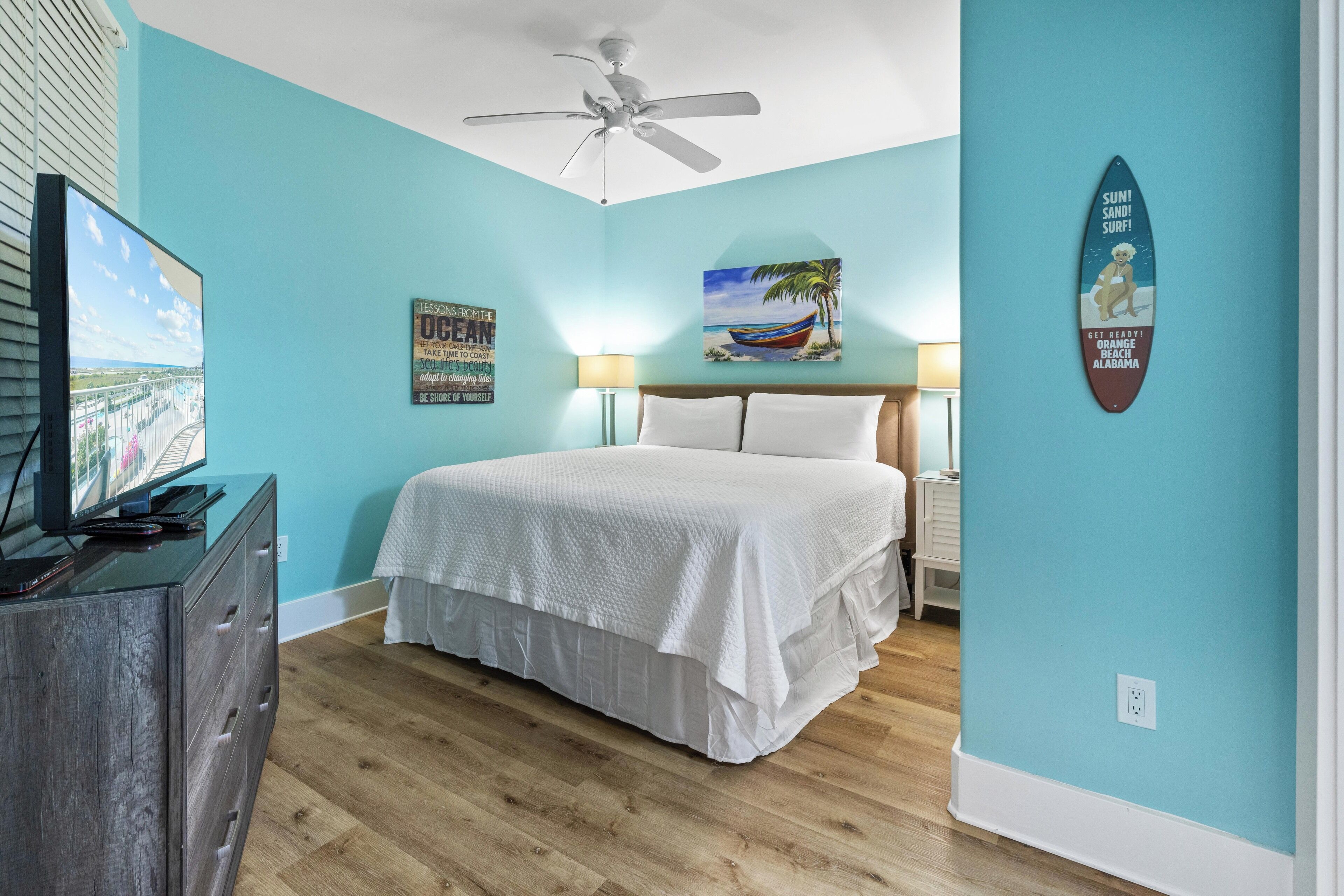 A bedroom with a bed, a ceiling fan, a TV, a dresser, and a surfboard decoration.
