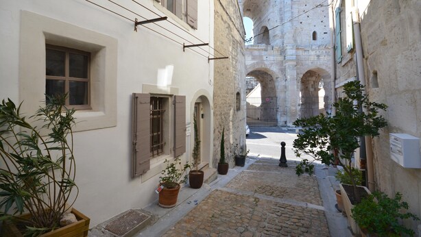 A narrow cobblestone street with buildings on either side, potted plants, and an arched stone structure in the background.