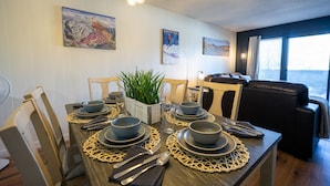 Een eetgedeelte met een houten tafel voor vier personen, omringd door stoelen. Er hangen foto’s in lijsten aan de muur, een kamerplant en een raam met zonwering.