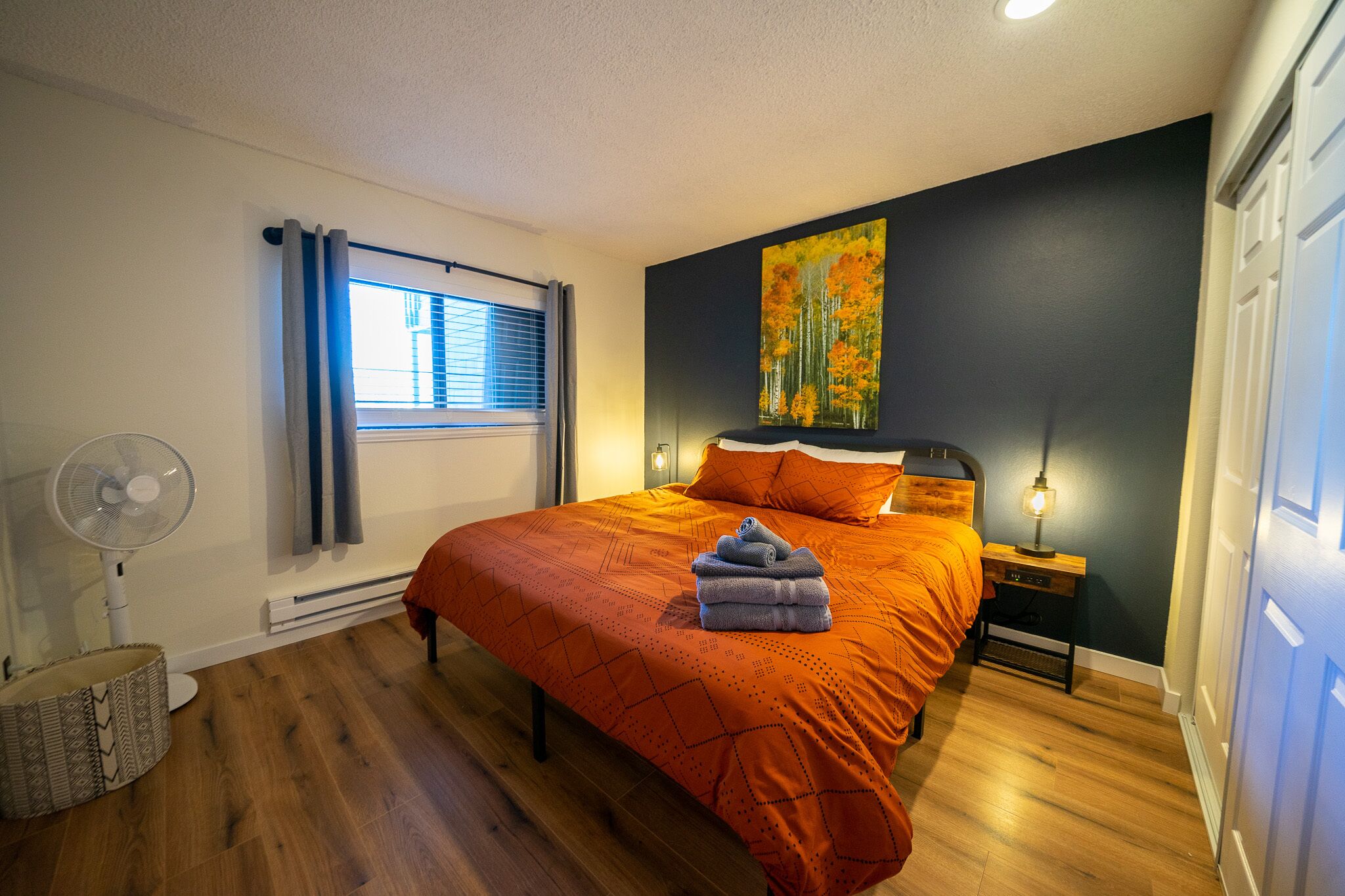 A bedroom with a bed, a standing fan, a heater, a nightstand with a lamp, and a window with curtains.