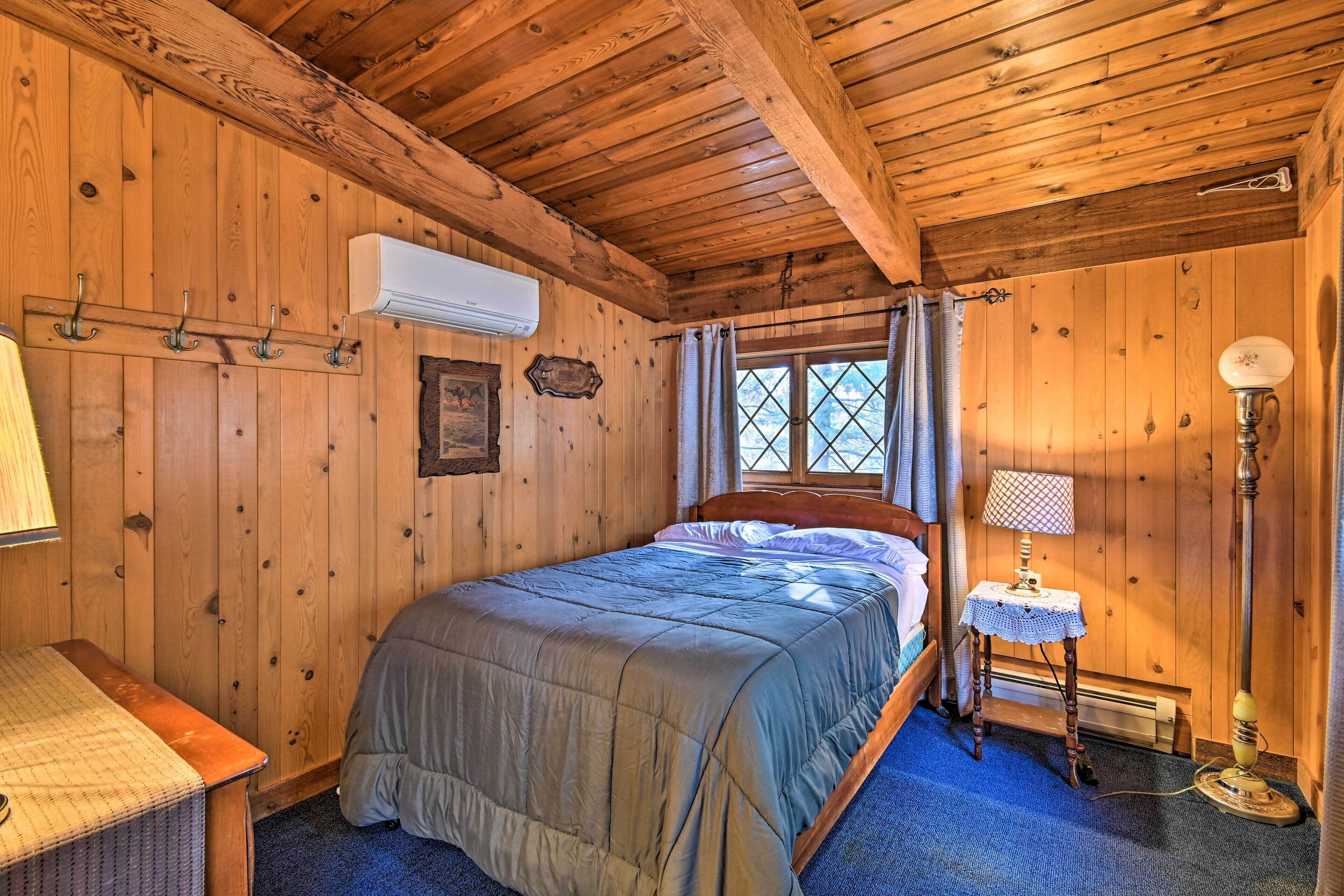 A wooden cabin bedroom with a bed, a small table with a lamp, and a window with curtains.