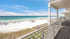 Blick auf einen Strand von einem Balkon mit Holzdeck und weißen Geländern, bewölkter, klarer Himmel.