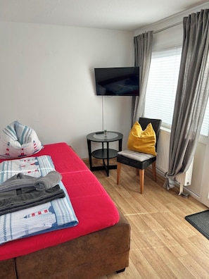 Ein Zimmer mit einem Bett, einem Fernseher, einem kleinen Tisch, einem Sessel und einem Fenster mit Vorhängen.