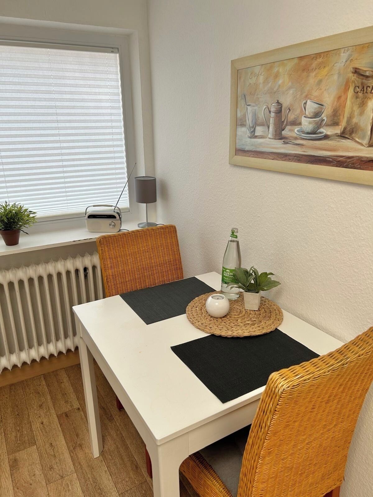 Ein Essbereich mit einem weißen Tisch, Korbstühlen, einer Heizung, einem Blumentopf, einem Radio und einem Bild an der Wand.