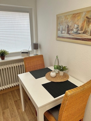 Ein Essbereich mit einem weißen Tisch, Korbstühlen, einer Heizung, einem Blumentopf, einem Radio und einem Bild an der Wand.