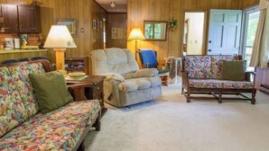 Ein gemütliches Wohnzimmer mit einer Couch, einem Sessel und einem Stuhl mit Blumenmuster. Es gibt einen Holzschrank und eine Lampe auf einem Tisch.