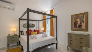 Un lit à baldaquin avec du linge de lit blanc, une table de chevet avec une lampe, une commode avec des tiroirs et un tableau encadré accroché au mur.