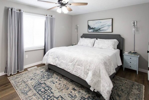 A bedroom with a bed, a ceiling fan, a bedside table, a rug, and a window with curtains.