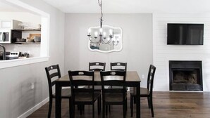 A dining area with a rectangular table and six chairs, a chandelier, a flat-screen TV mounted above a fireplace, and a kitchen area with cabinets and a microwave. - *NEW* Cozy, Smart Condo| 10 mins to DT, Biltmore (Asheville)