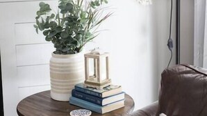 A cozy living room with a leather sofa, a small round table with a plant, books, and a lantern. A macrame wall hanging is visible. - *NEW* Cozy, Smart Condo| 10 mins to DT, Biltmore (Asheville)