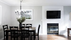A dining area with a wooden table and chairs, a chandelier, and a TV mounted above a fireplace. - *Comfy Smart Condo|10 mins to DT & (Asheville)