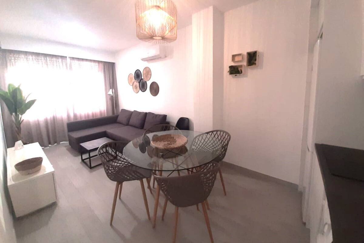 A modern living room with a glass dining table, two chairs, a sofa, and a large window with curtains.