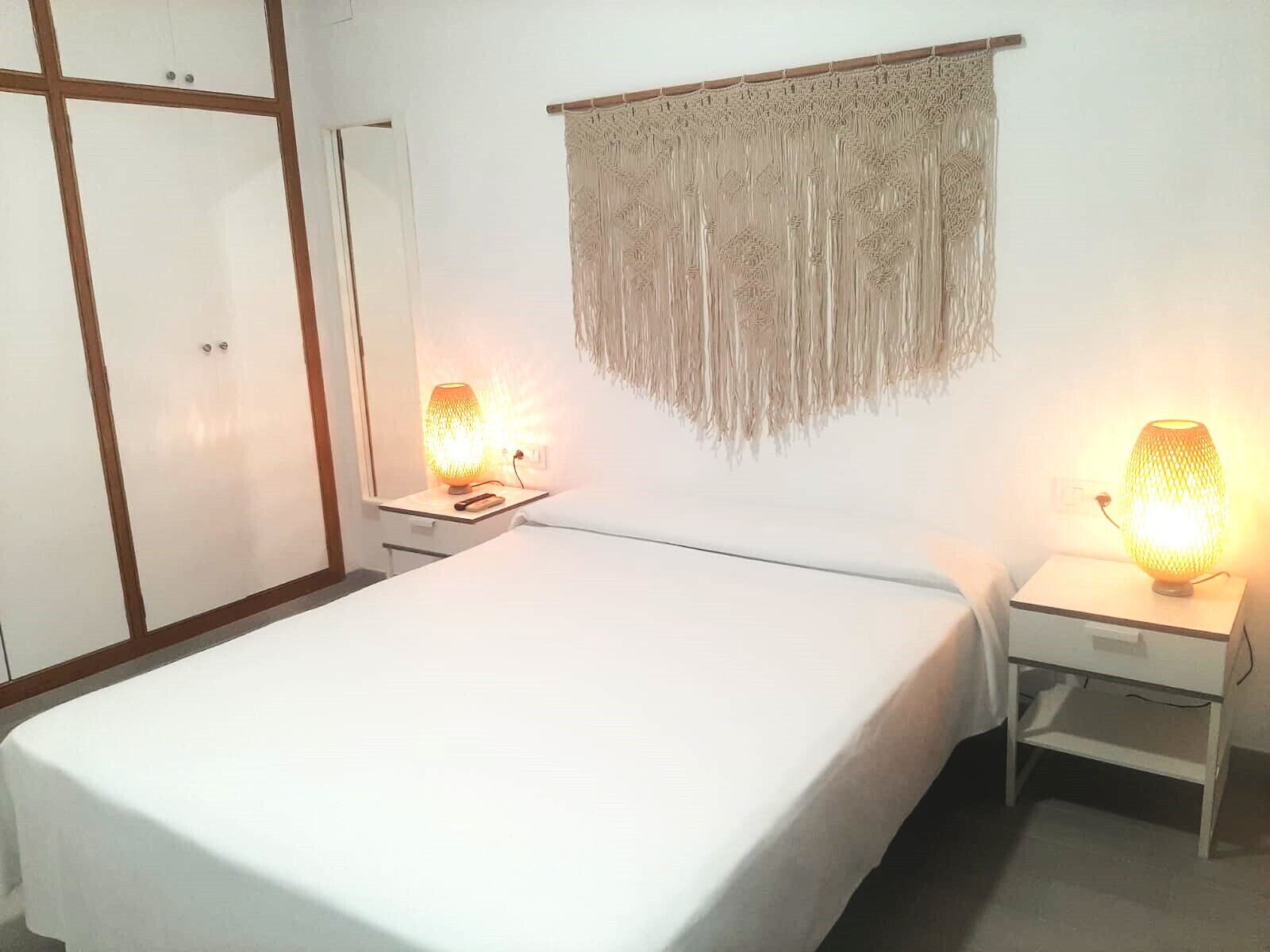 A bedroom with a bed, two bedside tables with lamps, a wardrobe, and a macramé wall hanging.