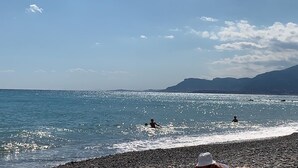 Ein Strand mit klarem, blauem Wasser, felsigem Ufer und Bergen im Hintergrund.