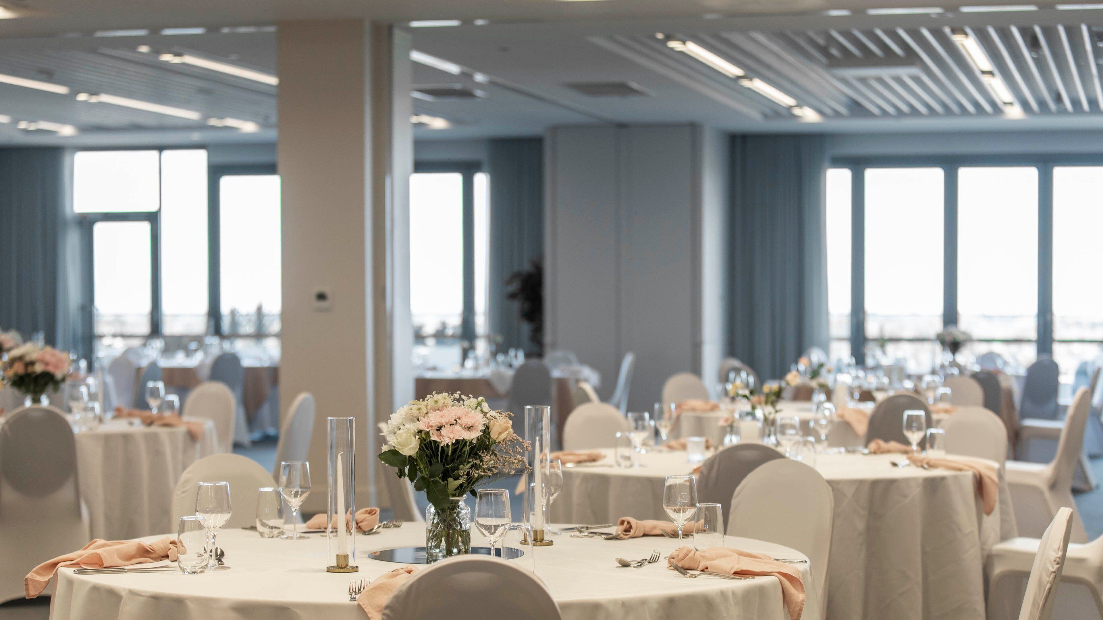 A banquet hall with round tables set for dining, each with a vase of flowers and candles.