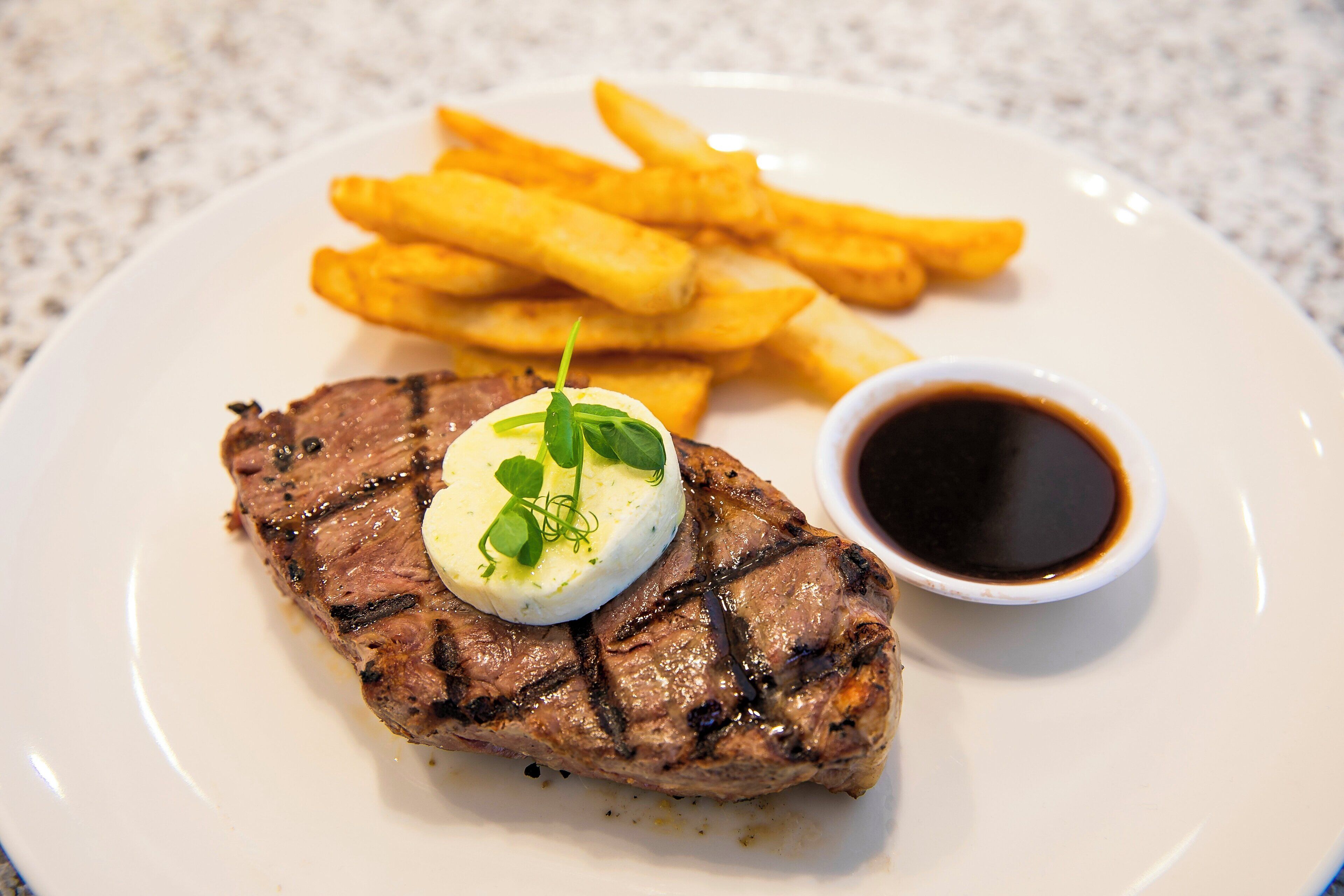 Un plato con un bistec a la parrilla, una guarnición de papas fritas y un recipiente pequeño de salsa.