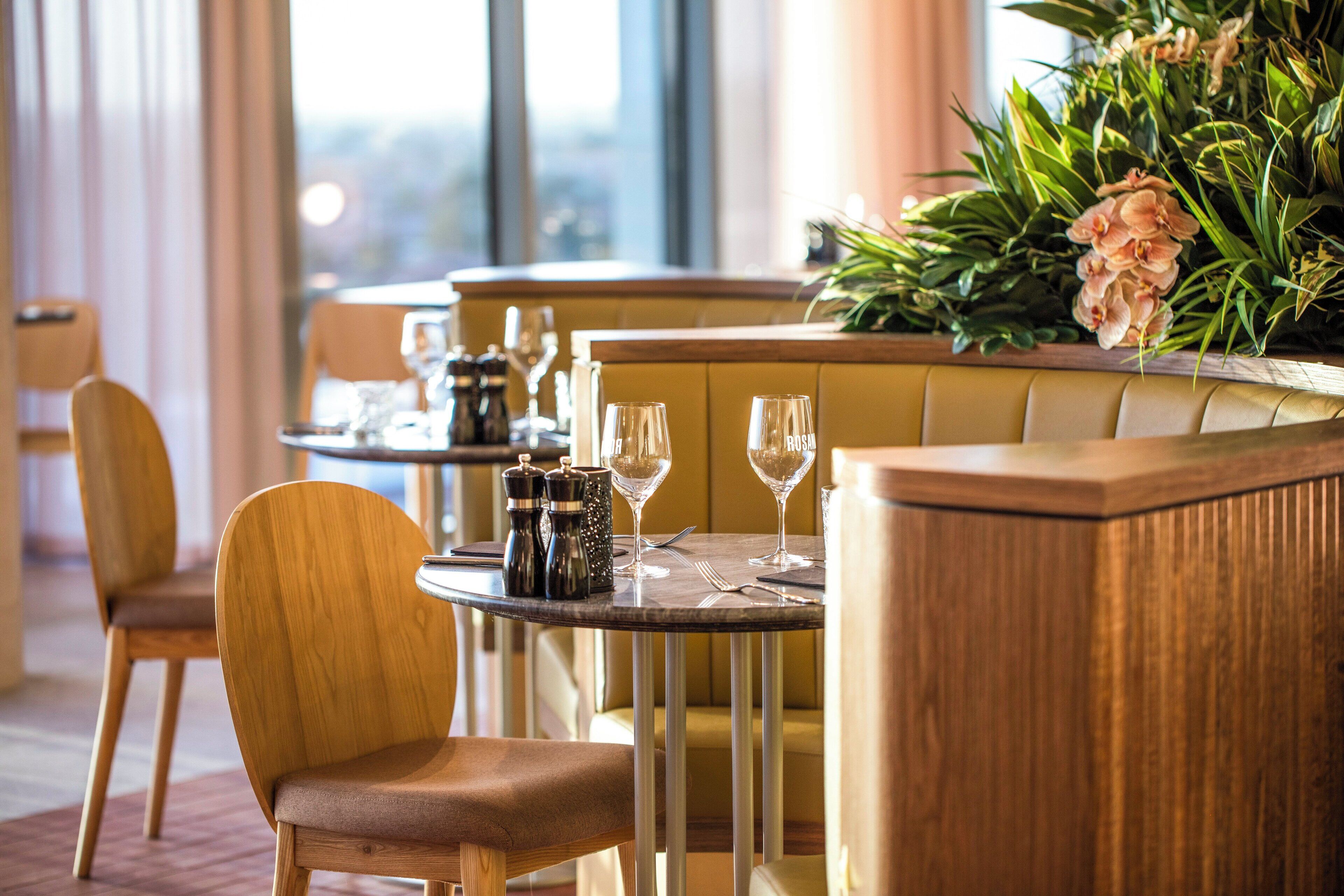 Ein Restaurantinneres mit Holztischen, gedeckten Tischen mit Weingläsern und einer dekorativen Pflanzengruppe.