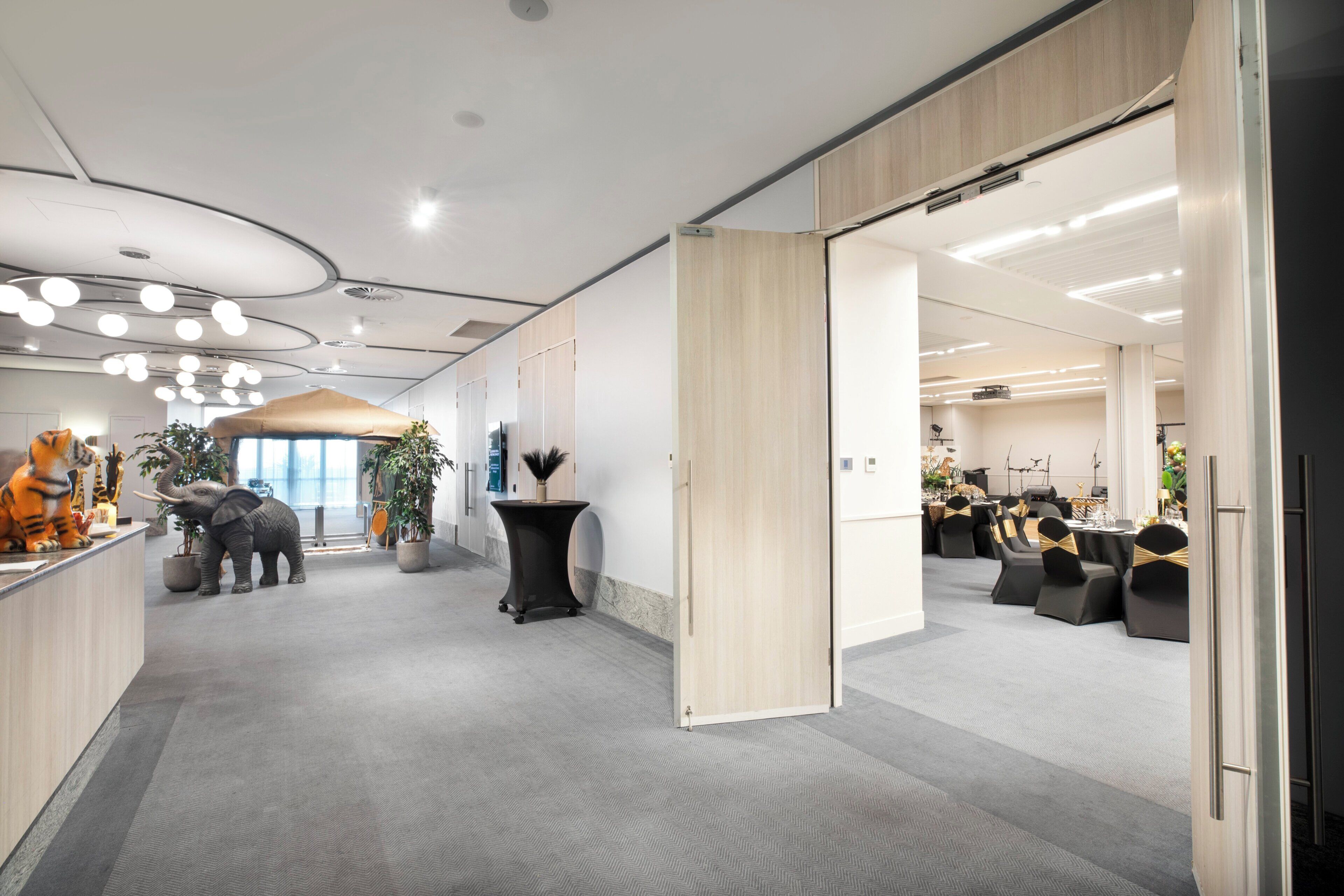 Un hall d’hôtel moderne avec un comptoir d’accueil, une grande plante en pot et un espace salon comprenant des tables et des chaises.
