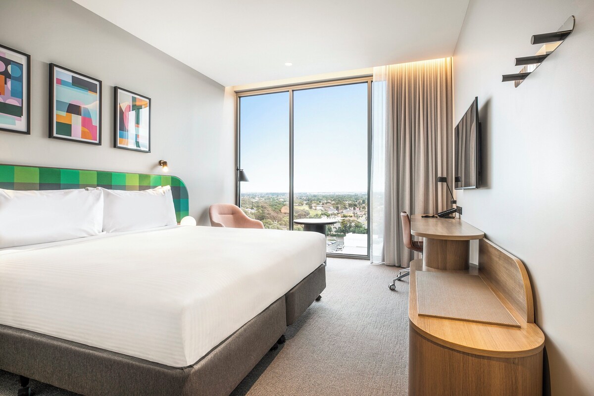 A hotel room with a large bed, a desk, a chair, a TV, and a view of the city.