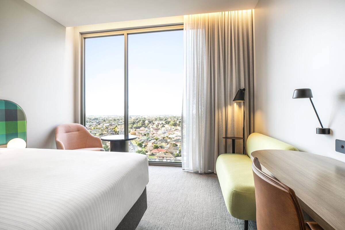 Habitación de hotel con una cama grande, un escritorio, una silla y vistas a un paisaje urbano a través de un ventanal.
