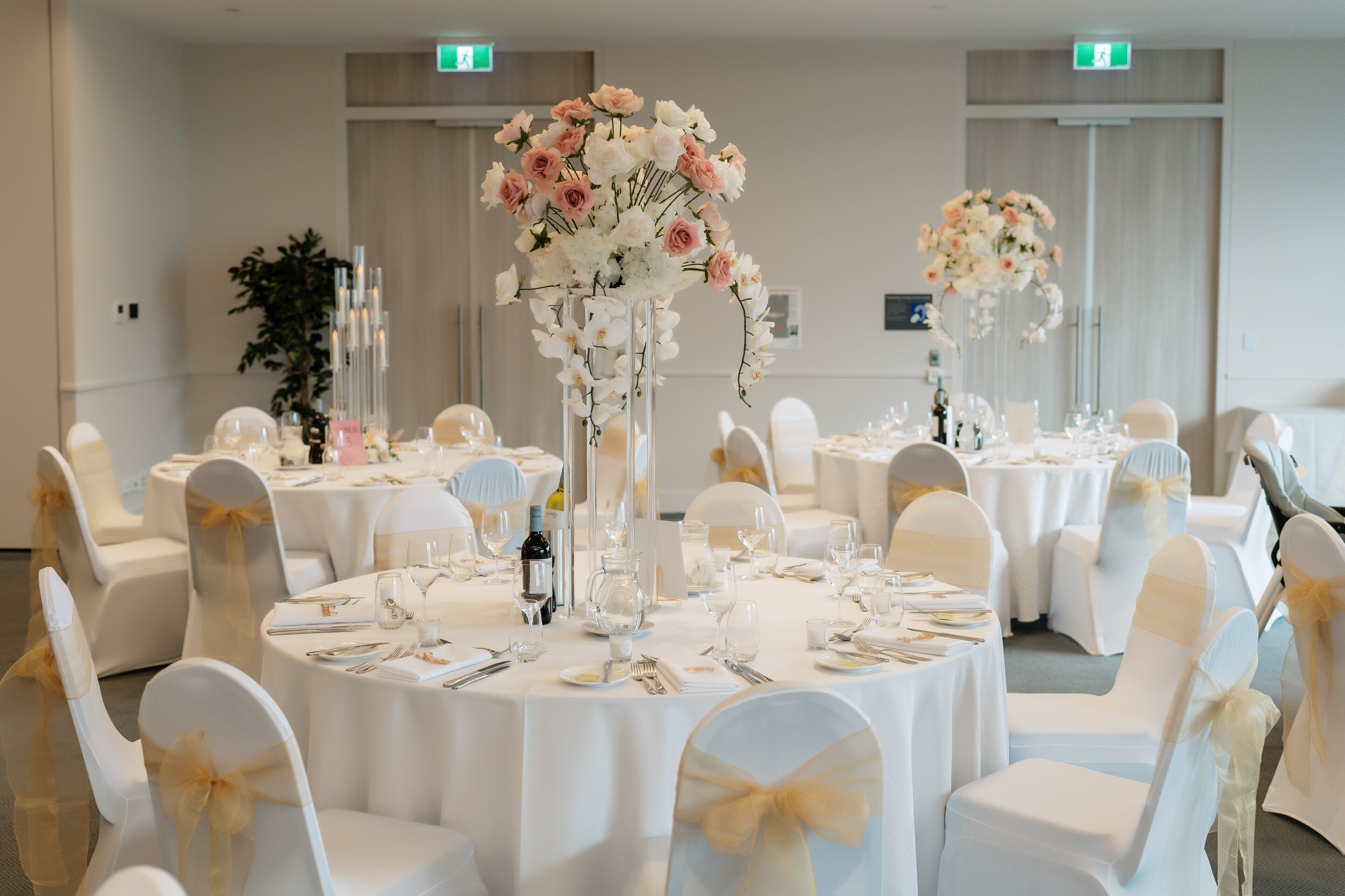 Un salón de banquetes con mesas redondas, sillas blancas con cintas doradas y centros de mesa florales.