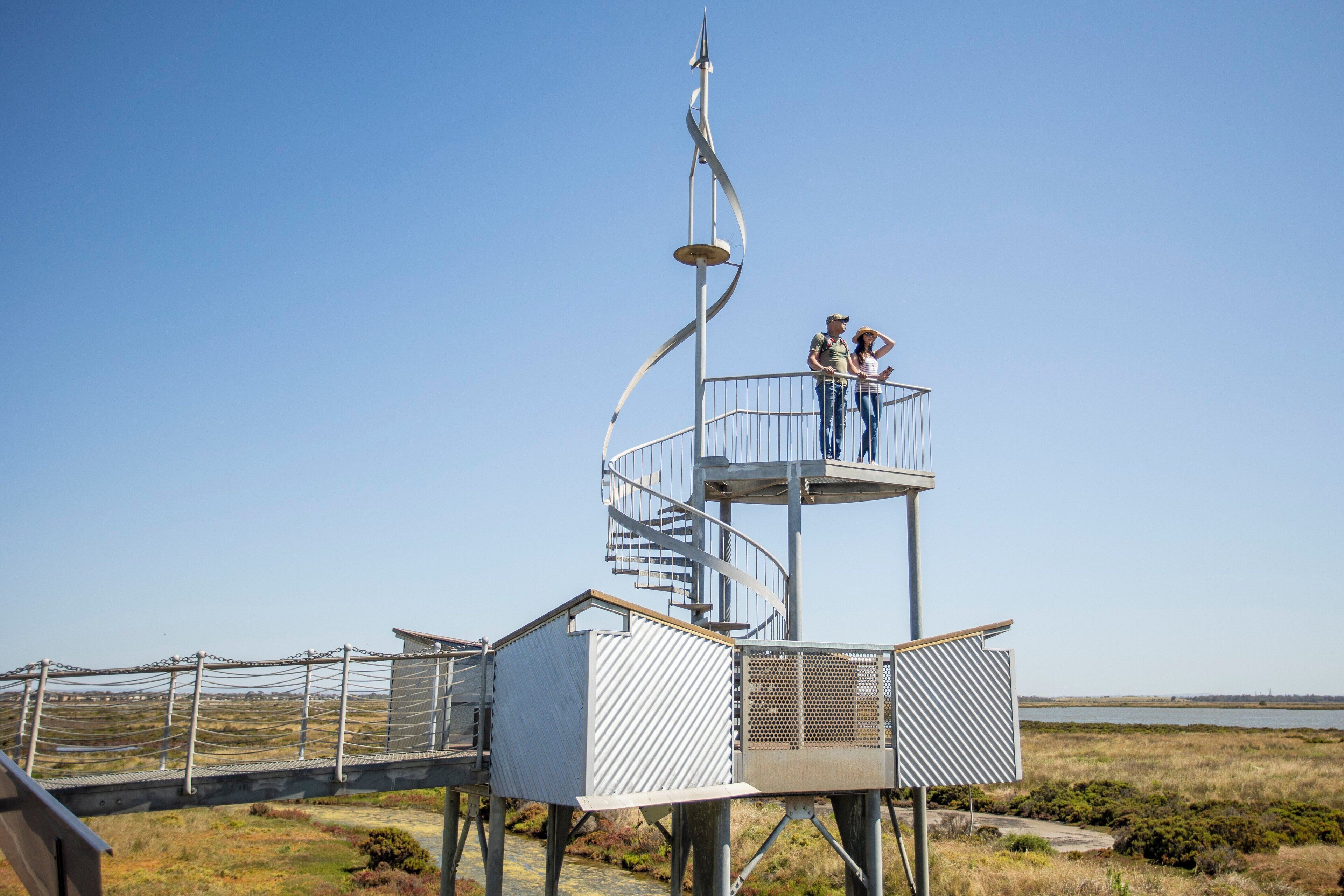 A spiral staircase leading to a viewing platform with a metal railing, overlooking a coastal landscape.