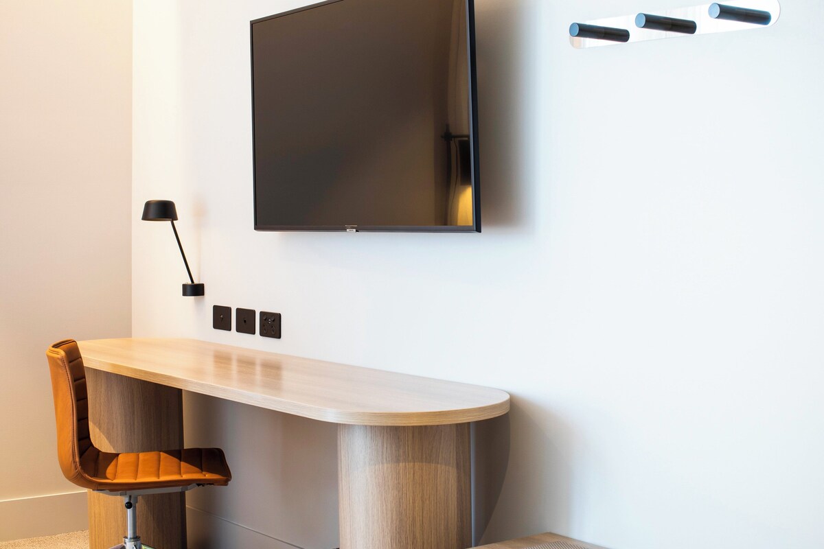 A modern room with a flat-screen TV mounted on the wall, a wooden desk with a chair, and a black desk lamp.