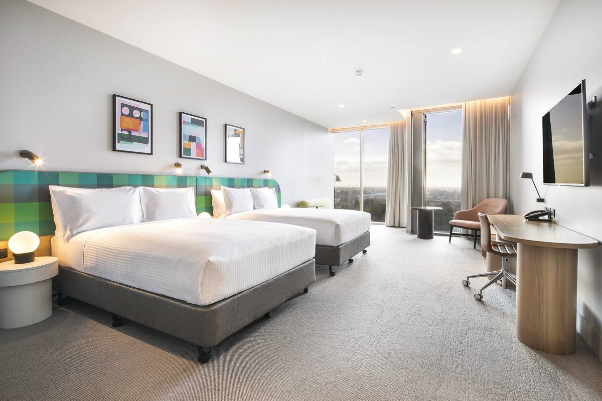 Habitación de hotel moderna con dos camas, un escritorio, una silla y un ventanal con vistas a la ciudad.