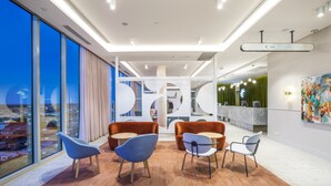 Um lobby moderno com uma grande parede de vidro, área de estar com cadeiras, balcão de recepção e placa indicando os sanitários.